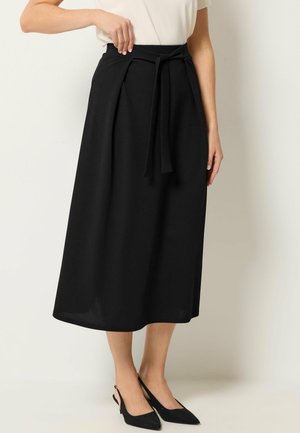 Kvinde iført sort midinederdel med høj talje og bindebælte, kombineret med sorte slingback-hæle med spids tå og hvid top.