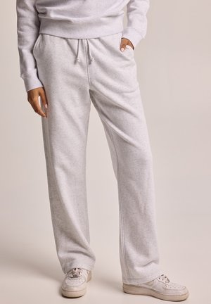 Persoon draagt lichtgrijze joggingbroek met trekkoord in de taille, witte sneakers en een bijpassende lichtgrijze trui met handen in de zakken.