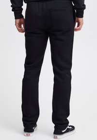 Juodi sportinės kelnės su šiek tiek siaurėjančia apačia, pagamintos iš minkštos medžiagos, su užpakaliniu kišeniu, derinamos su juodais sportiniais batais.