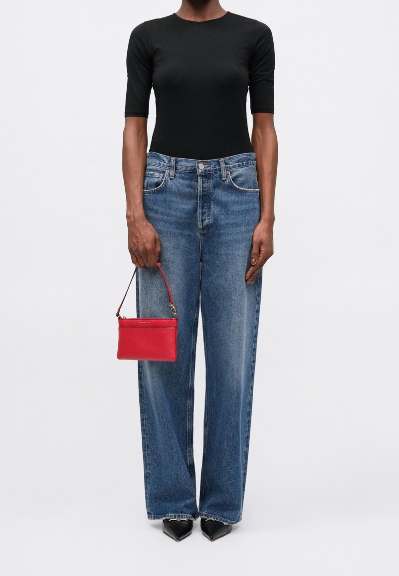 Personne portant un haut noir ajusté, un jean bleu à jambes larges, des chaussures noires pointues et tenant un petit sac à main rouge à ses côtés.