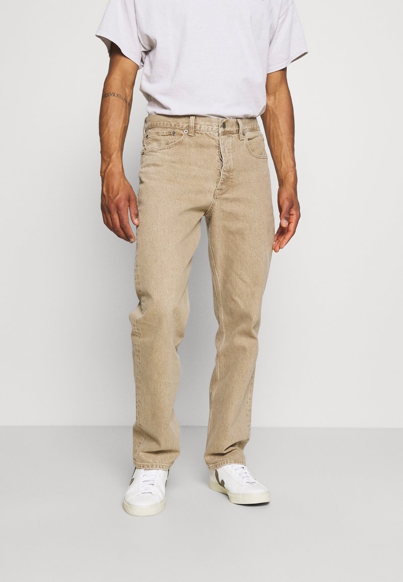 Jeans in denim beige con una vestibilità dritta, dotati di chiusura frontale con bottone e zip, cinque tasche e una leggera scoloritura. Abbinati a sneakers bianche.