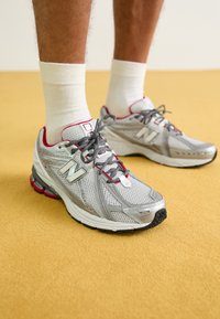 Szare siatkowe buty sportowe z czerwonymi akcentami, teksturowanymi nakładkami i szarymi sznurówkami. Grube, wyprofilowane podeszwy zapewniają wsparcie i trwałość.