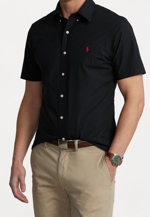 Mann trägt ein schwarzes kurzärmeliges Hemd mit rotem Logo und beiger Hose, Hand in der Tasche und braune Uhr am linken Handgelenk.