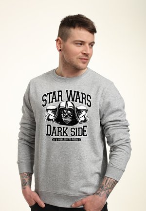 Mand med tatoveringer iført grå sweatshirt med Star Wars Dark Side grafik og tekst, kigger til siden med hænderne i lommerne.