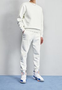 Man i vit sweatshirt och joggers med blå och orange hög-topp sneakers, som står på en blå mattad golv framför en vit vägg.