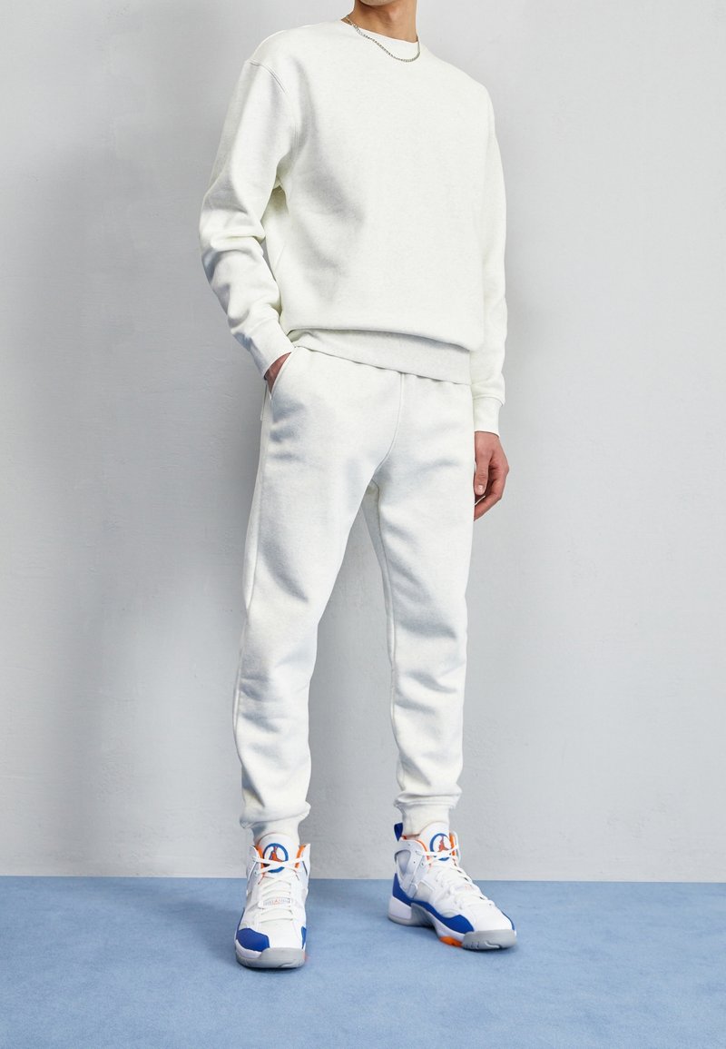 Man i vit sweatshirt och joggers med blå och orange hög-topp sneakers, som står på en blå mattad golv framför en vit vägg.