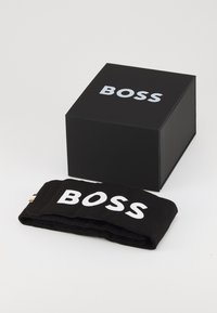 Juodas audinio apyrankė su baltu "BOSS" logotipu, padėta šalia juodo dėžutės, ant kurios viršaus aiškiai matomas tas pats logotipas.