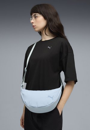 Lichtblauwe schoudertas met een gebogen ontwerp, voorzien van een gladde textuur en geborduurd logo. Gecombineerd met een zwart T-shirt en verstelbare band.