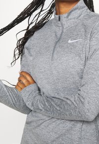 Grijze ritsathletische top van een textuurstof. Heeft een hoge kraag en lange mouwen, met een klein wit Nike-logo op de borst.