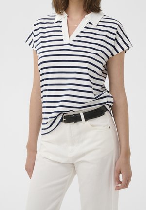 Vrouw draagt een wit shirt met korte mouwen met marineblauwe horizontale strepen en een witte kraag, ingestopt in een witte broek met een zwarte riem.