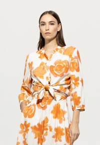 Bluse mit Blumenmuster und orangefarbenen Rosen auf weißem Hintergrund. Mit geknoteter Vorderseite, Knopfverschluss und Dreiviertelärmeln.