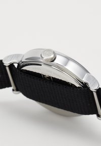 Orologio in metallo argento con cassa rotonda, cinturino in tessuto nero e corona testurizzata contrassegnata con "We". Resistente all'acqua fino a 50 metri.