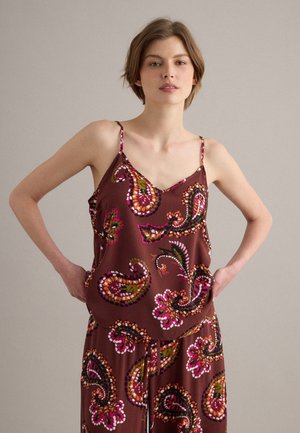 Femme portant un haut à fines bretelles marron à motif paisley et un pantalon assorti, debout les mains sur les hanches devant un fond uni.