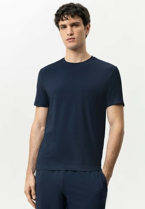 Junger Mann mit lockigem dunklem Haar, der ein schlichtes marineblaues T-Shirt mit kurzen Ärmeln und passende Hosen vor hellem Hintergrund trägt.