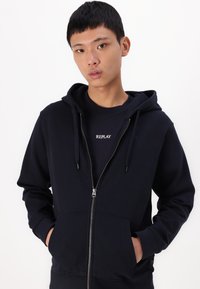 Mornarska zip-up kapuca z notranjim žepom, kapuco na vrvico in rebrastimi manžetami. Nošena čez črno majico z belim logotipom "REPLAY".