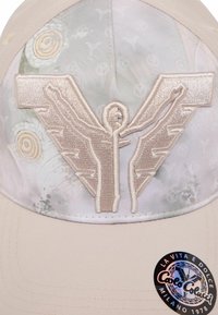 Beige Kappe mit besticktem Logo, das stilisierte Flügel zeigt, strukturiertem Stoff mit floralen Mustern und einem kreisförmigen Markenaufkleber an der Seite.