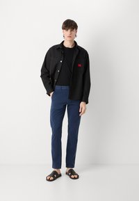 Cămașă neagră cu nasturi, având două buzunare frontale, asortată cu pantaloni bleumarin și sandale negre cu talpă plată, pe un fundal gri deschis.