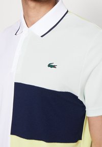 Herrpolo med ett design av vit, ljusgrön och marinblå färgblock. Har en klassisk krage, två knappar och en grön krokodil-logotyp.