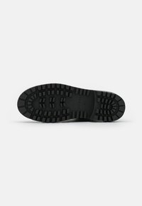 Semelle en caoutchouc noir avec un motif de grip texturé, comportant des crampons surélevés pour l'adhérence. Logo de la marque visible sur la semelle intermédiaire.