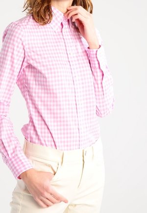 Kobieta w różowo-białej koszuli w kratę zapiętej na guziki, wpuszczonej w kremowe spodnie, jedna ręka przy kołnierzu, druga w kieszeni.
