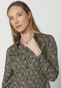 Camicia nera con bottoni, decorata con un motivo floreale in giallo e bianco. Presenta un colletto e maniche lunghe con polsini arricciati. Tessuto dalla consistenza liscia.