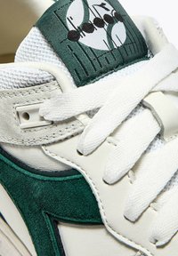 Sneaker bianco con dettagli in suede verde, linguetta in rete e chiusura con lacci. Presenta un logo distintivo sulla linguetta e dettagli di cucitura in contrasto.