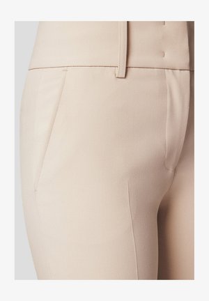 Beżowe spodnie dopasowane o gładkiej fakturze, z boczną kieszenią i bezpiecznym pasem. Tkanina wydaje się lekka i dobrze ustrukturyzowana.