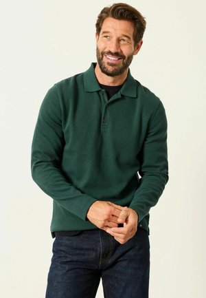 Mann mit Bart, der lächelt, trägt ein dunkelgrünes langärmeliges Polohemd und dunkle Jeans, steht vor einem schlichten hellen Hintergrund.