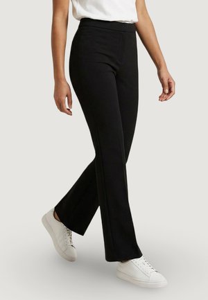 Persona che cammina, indossa pantaloni neri a gamba dritta, sneaker bianche e una maglietta bianca a maniche corte su uno sfondo uniforme.