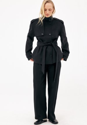 Kobieta w czarnym wełnianym płaszczu z paskiem i pagonami na ramionach, czarnych szerokich spodniach i czarnych butach, stojąca na tle jednolitej białej ściany.