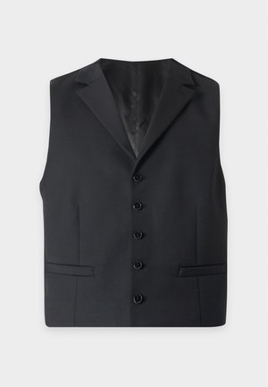 Homme portant une chemise blanche à manches courtes, un gilet noir boutonné, un pantalon noir assorti et des chaussures habillées noires, debout les mains dans les poches.