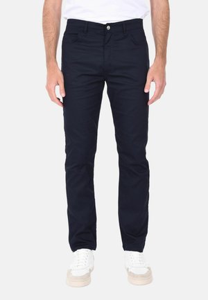 Uomo che indossa jeans neri slim fit e sneakers bianche, in piedi con le mani rilassate lungo i fianchi su uno sfondo bianco neutro.