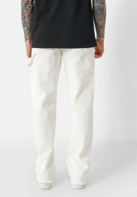 Weiße Cargo-Hosen mit lockerer Passform, ausgestattet mit Seitentaschen, fransigen Säumen und leichtem Stoff. Getragen mit einem schwarzen Shirt.