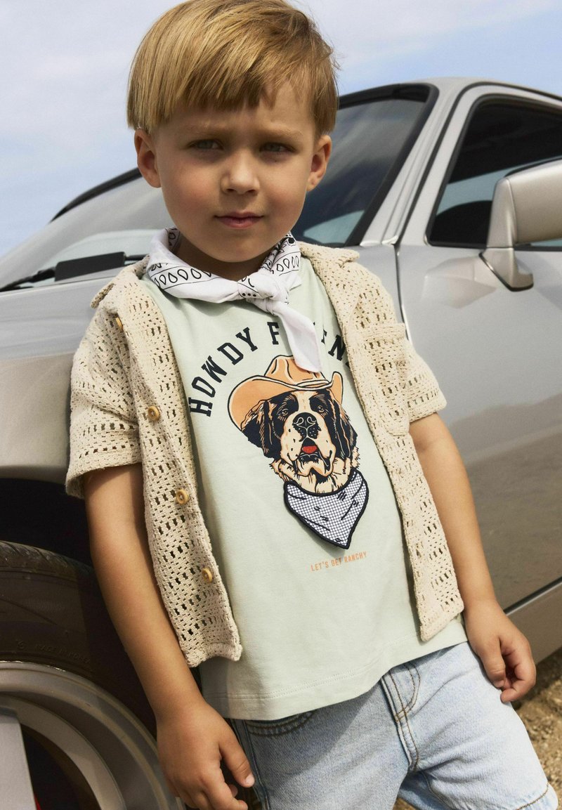 Bambino che indossa una maglietta verde chiaro con una grafica di un cane, un cardigan di maglia beige, un bandana bianco e pantaloni di jeans, posato vicino a un'auto.