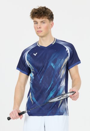 Ung mand, der holder en badmintonketcher iført en blå mønstret sportstrøje og hvide shorts mod en ensfarvet baggrund.