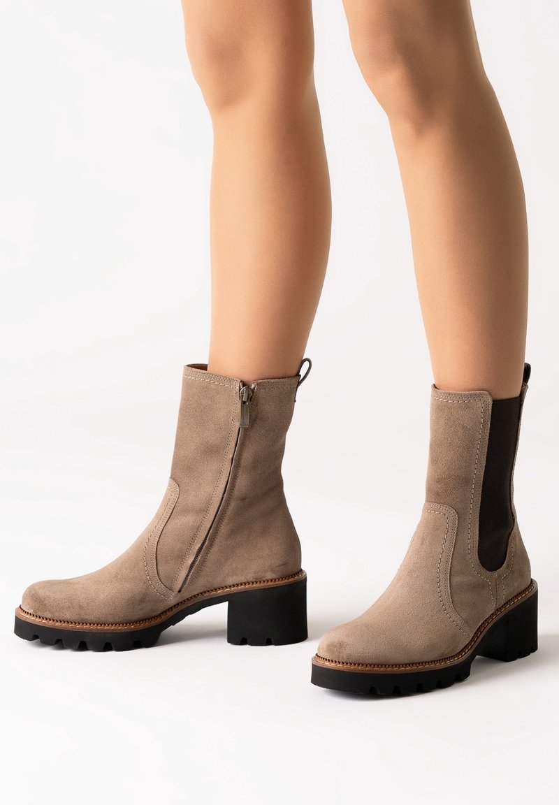 Bottes taupe en daim montant à la cheville avec talons épais noirs, fermeture éclair latérale sur une botte, panneau élastique sur l'autre, portées sur jambes nues.