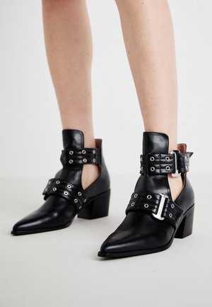 Černé kožené kotníkové boty s špičatými špičkami, vybavené úchytkami a dvěma nastavovacími přezkami. Blokové podpatky zajišťují stabilitu.