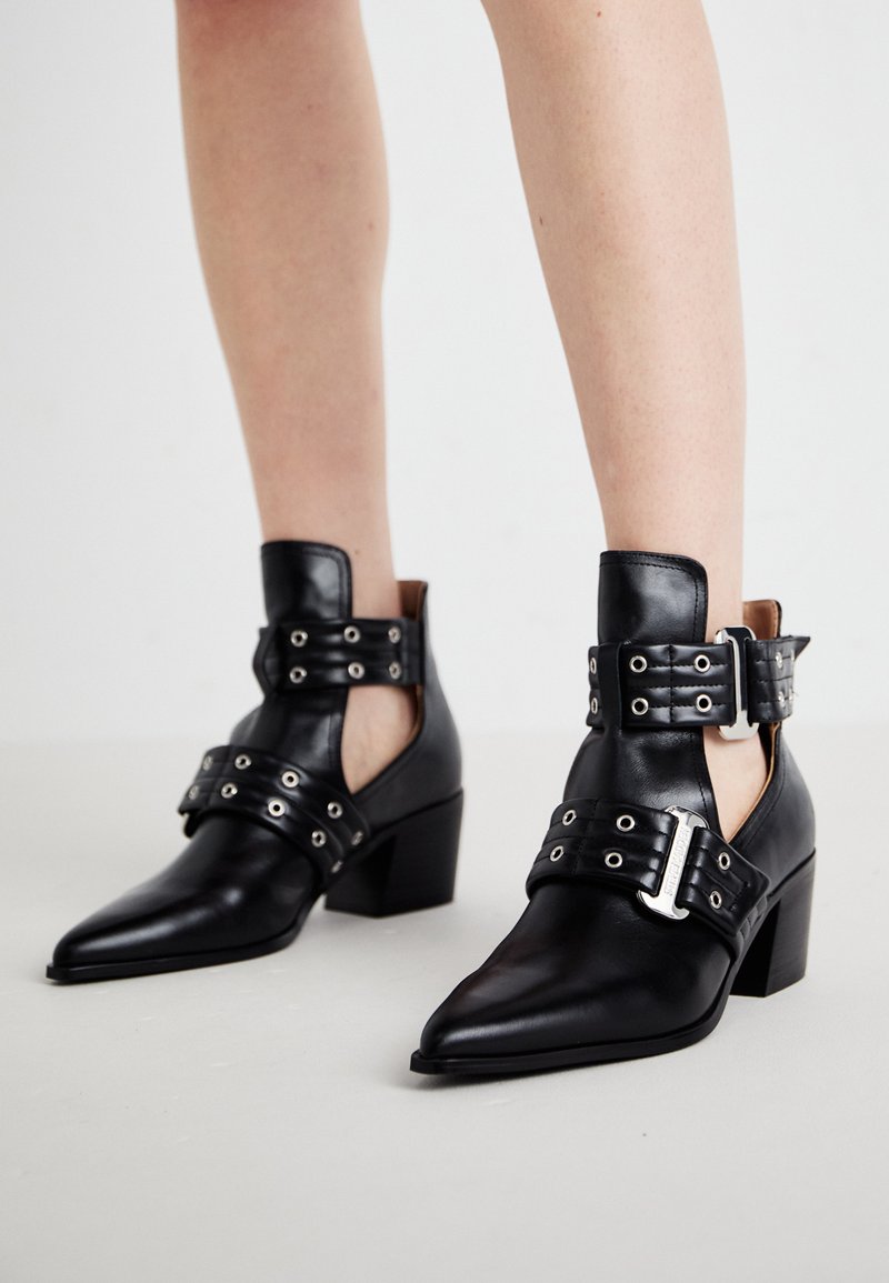 Stivali neri in pelle fino alla caviglia con punte appuntite, dotati di occhielli e due cinturini regolabili con fibbie. I tacchi a blocco offrono stabilità.