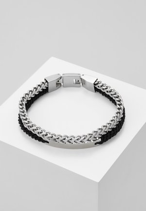 Silver chain bracelet with black braided cord and clasp, displayed on a white angled pedestal against a gray background.