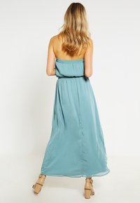 Femme aux cheveux blonds portant une robe maxi sans bretelles de couleur bleu clair et des sandales à talons beiges, se tenant de dos contre un fond blanc.