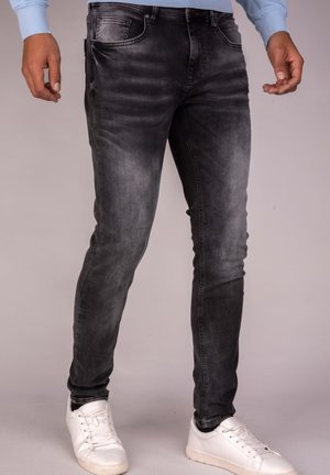 Mężczyzna ubrany w obcisłe, wyblakłe czarne dżinsy i białe trampki, stojący na tle gładkiego jasnego tła.