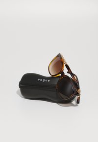 Sunglassi iz želve školjke z gradijentnimi lečami, z zlati naglasi na templjih, počivajo na črnem zadrgo zaprtem etuiju z logotipom "vogue".