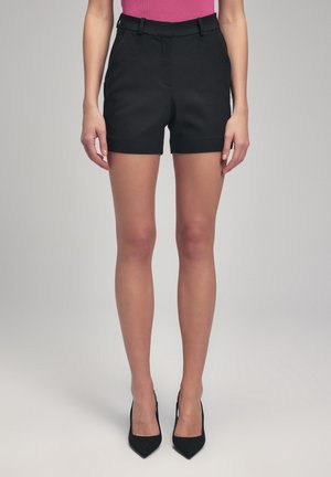 Femme portant un short taille haute noir ajusté avec passants de ceinture et des escarpins noirs à bout pointu sur un fond neutre.