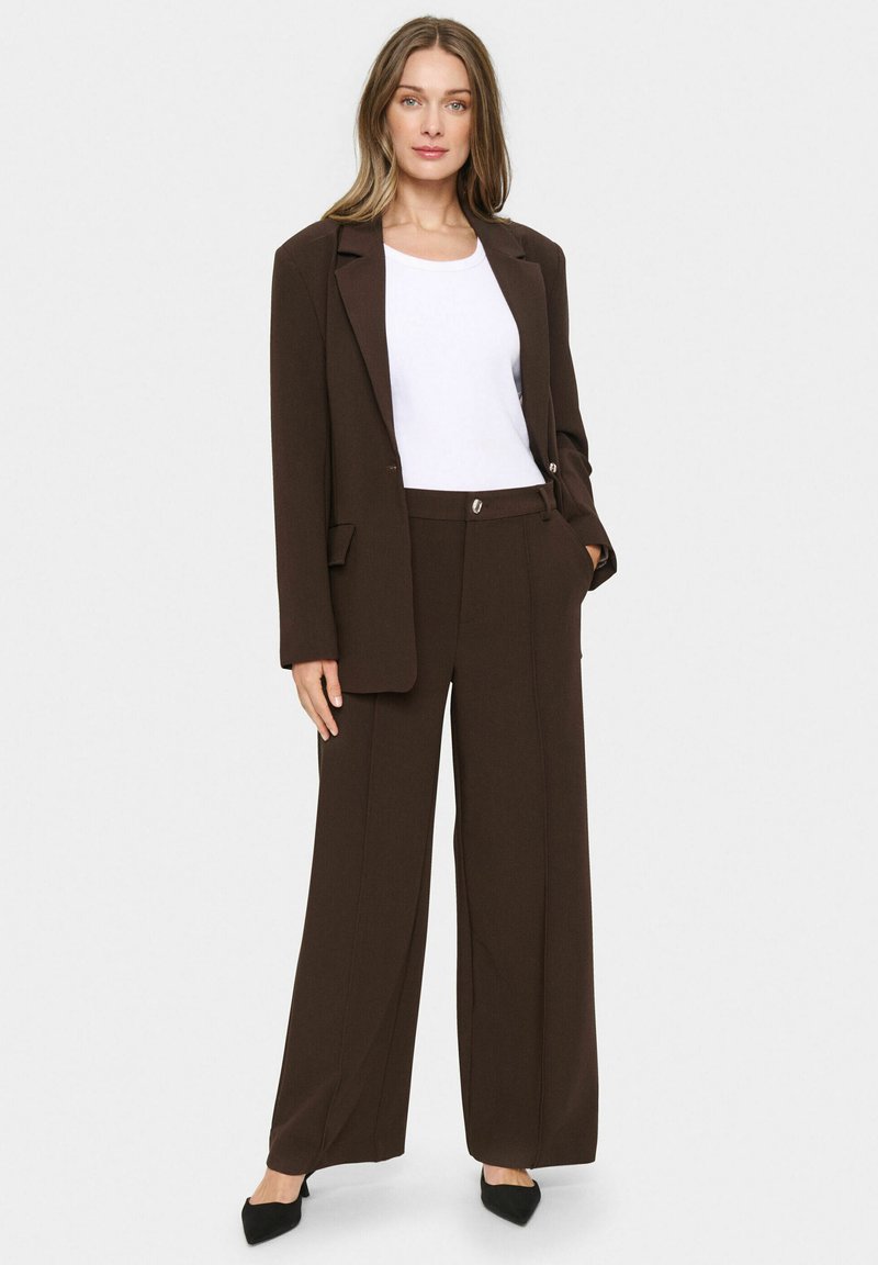 Brązowa dopasowana marynarka z szerokimi spodniami w tym samym kolorze, charakteryzująca się eleganckim wzornictwem i dyskretnym połyskiem. Biała dopasowana bluzka pod spodem. Czarne czółenka z ostrym noskiem.