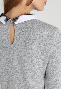 Grå tröja med texturerad stickning, med en vit krage och svart fransdetalj på baksidan, samt en nyckelhålsöppning vid halsen.