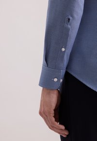 Man in een blauw gemusterd overhemd met lange mouwen en knoopmanchetten, en zwarte broek, met een ontspannen hand naast zich tegen een effen achtergrond.