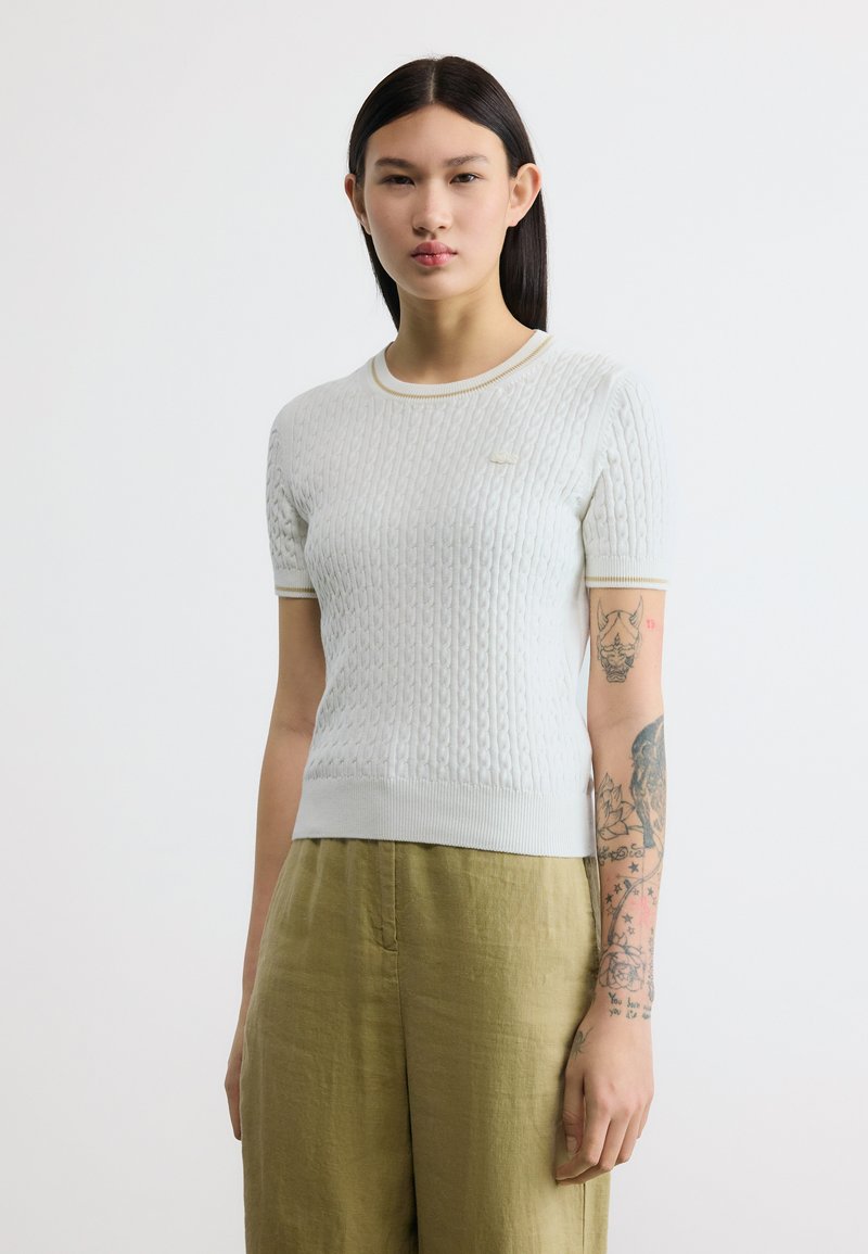 Νεαρή γυναίκα με ίσια σκουρόχρωμα μαλλιά, που φοράει λευκό πλεκτό τοπ με κοντά μανίκια και ανοιχτοπράσινο παντελόνι, με ορατά τατουάζ στα χέρια.