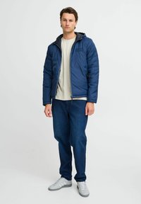 Marineblå puffjakke med hette, frontglidelås og elastiske mansjetter, kombinert med en lyse skjorte og mørke denimjeans, iført grå sneakers.