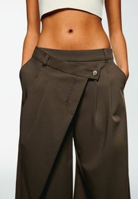 Pantaloni a gamba larga marroni con una vita strutturata, dotati di chiusura con bottone singolo e tasche frontali. Texture liscia con un aspetto sartoriale.