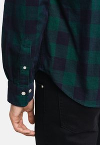 Groene en marineblauwe geruite corduroy overhemd met knoopmanschetten, met textielstof, zichtbare stiksels en een gebogen zoom, gedragen over zwarte broek.